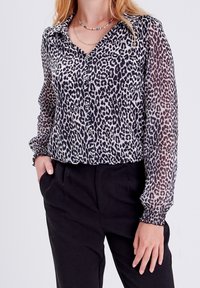 Leopardenmuster-Bluse aus transparenter Stoff mit Knopfleiste und Kragen. Sie hat lange, geraffte Ärmel und eine lockere Passform, kombiniert mit schwarzen Hosen.
