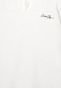 Polo bianco con un motivo a maglia testurizzato, dotato di scollo a V e un piccolo logo ricamato nero con la scritta "Crave Joy."