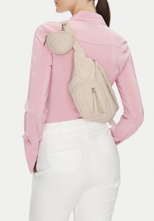 Kobieta nosząca pikowaną beżową torbę na ramię z wieloma zamykanymi na zamek kieszeniami, na różowej bluzce i białych spodniach.