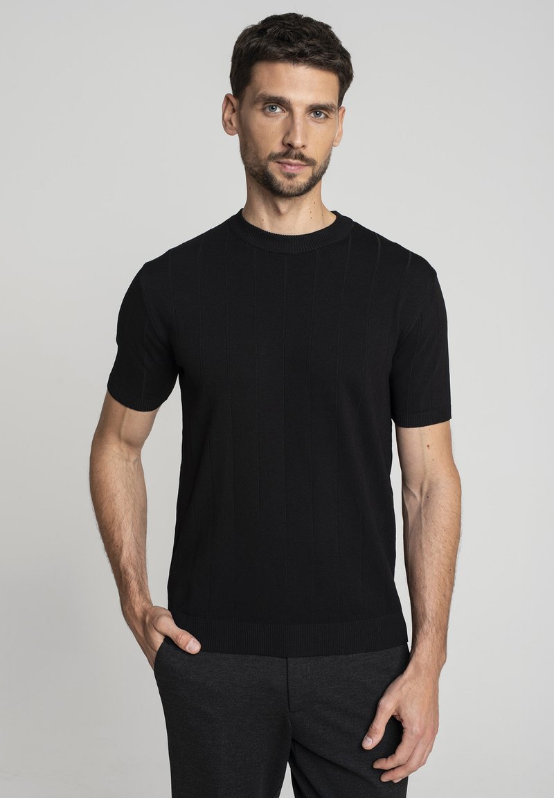 Mężczyzna z krótkimi ciemnymi włosami i brodą, ubrany w dopasowany czarny sweter z krótkim rękawem oraz ciemne spodnie, stojący z jedną ręką w kieszeni.