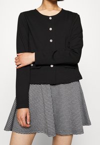 Černý krátký svetr s dlouhými rukávy a kulatým výstřihem, s kovovými stříbrnými knoflíky a dvěma předními kapsami, v kombinaci s černobílou kostkovanou sukní.