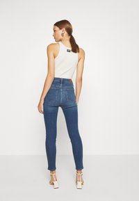Jeans skinny de cintura alta azuis com bolsos traseiros e tecido texturizado, combinados com uma blusa sem mangas branca e sandálias de bloco de salto creme.