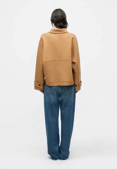 Personne de dos portant une veste couleur camel avec des poignets boutonnés et un jean bleu ample sur un fond blanc uni.