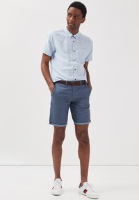 Ljusblå kortärmad skjorta med knappar och bröstficka, ihopparad med marinblå shorts med uppvikta kanter och ett mönstrat bälte. Vita sneakers.