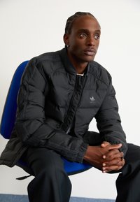 Schwarze Pufferjacke mit gerippten Bündchen, besticktem Logo und Reißverschluss, getragen über einem hellen Hemd, sitzend auf einem blauen Stuhl.