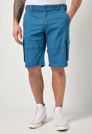 Mand iført blå cargoshorts og hvide høje sneakers, stående med den ene hånd i lommen og en synlig tatoveret arm.