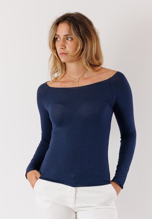 Maglietta navy a maniche lunghe con scollatura off-the-shoulder, vestibilità slim e texture liscia, abbinata a pantaloni chiari.