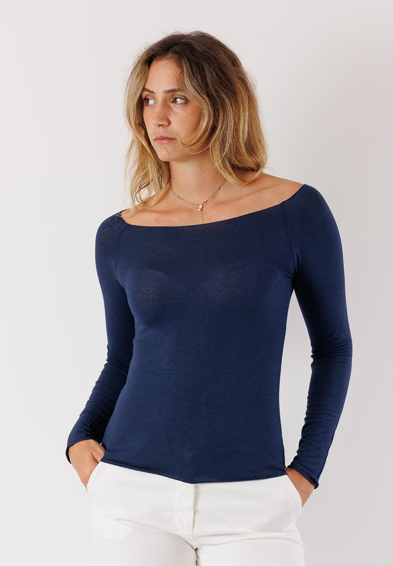 Maglietta navy a maniche lunghe con scollatura off-the-shoulder, vestibilità slim e texture liscia, abbinata a pantaloni chiari.