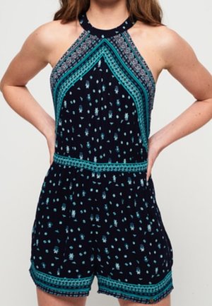 Femme portant une combinaison sans manches bleu marine avec des motifs géométriques et floraux turquoise et blancs, debout les mains sur les hanches.