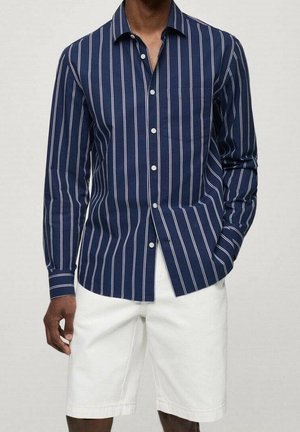 Mężczyzna w granatowej, długorękawowej koszuli na guziki z białymi pionowymi paskami i białych, sięgających do kolan szortach, z ręką w kieszeni.