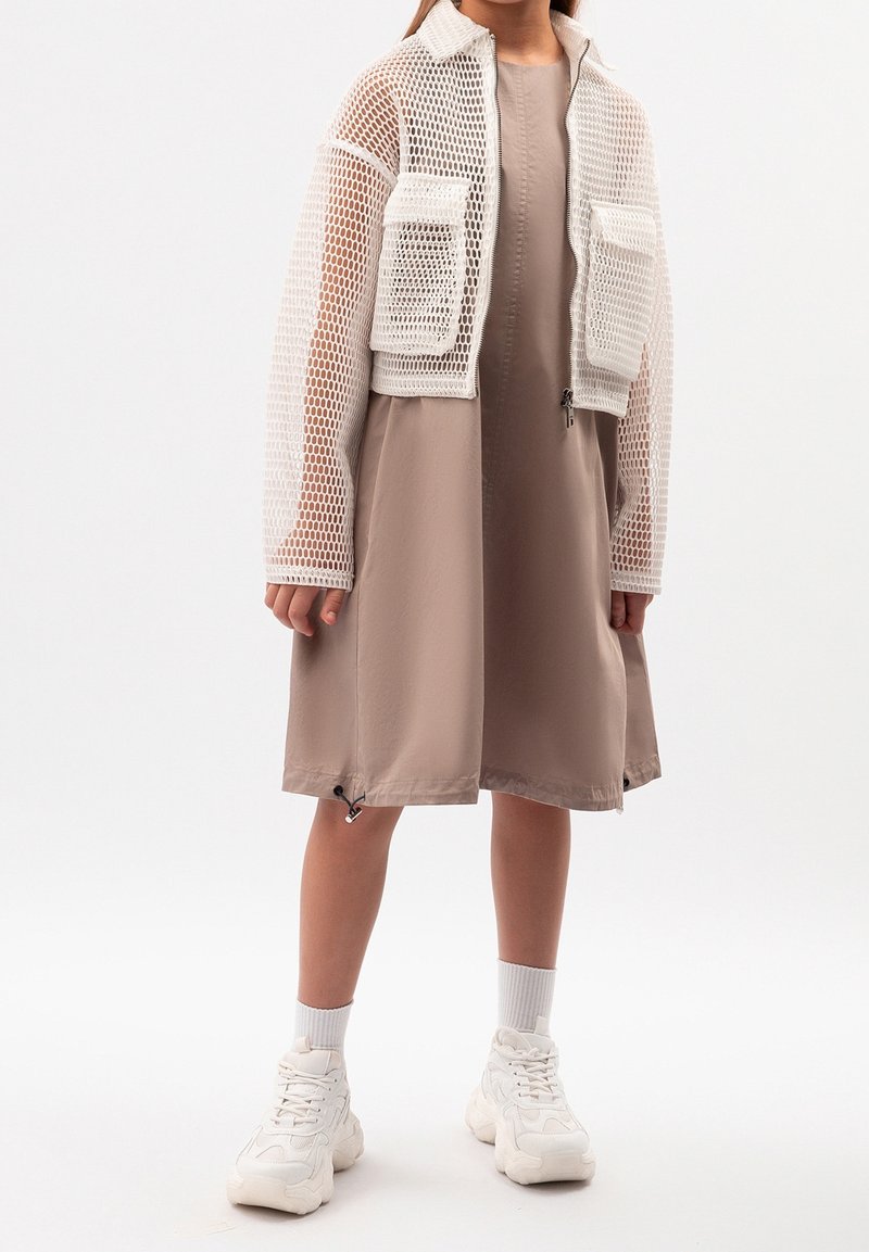 Veste blanche courte en résille avec deux poches avant portée sur une robe beige. Les chaussures sont des baskets blanches. La robe présente une texture lisse et une coupe décontractée.
