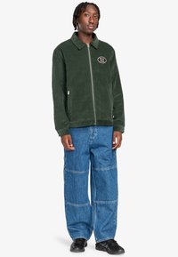 Grøn fløjl zip-up jakke med krave, prydet med et cirkulært logo patch. Kombineret med løstsiddende blå denim bukser og sorte sneakers.