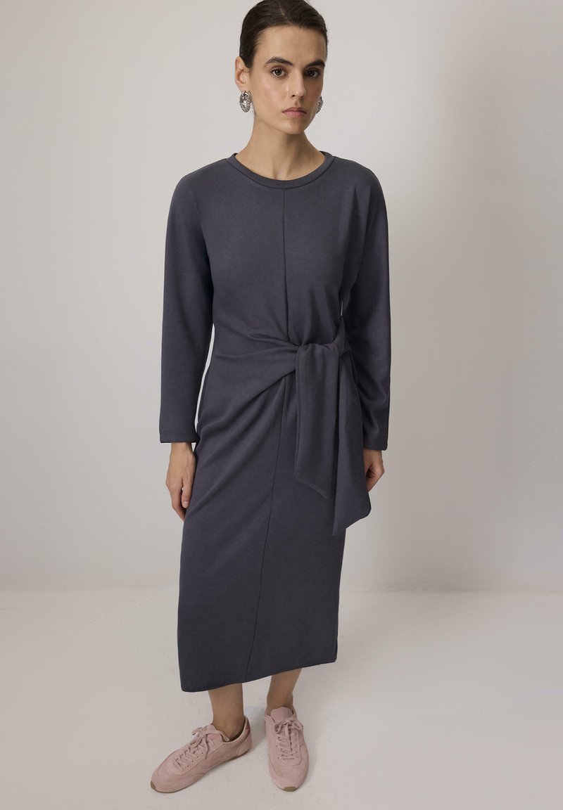 Vestido de manga larga en gris oscuro con un detalle de cintura atada. Textura suave, hasta la rodilla, combinado con zapatillas de deporte rosa claro. Diseño simple y ajustado.