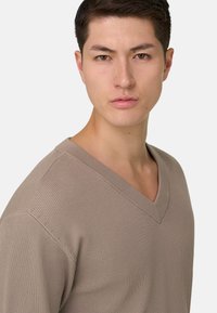 Jeune homme aux cheveux courts et foncés, portant un pull en maille beige à col en V, regardant droit devant avec une expression neutre.