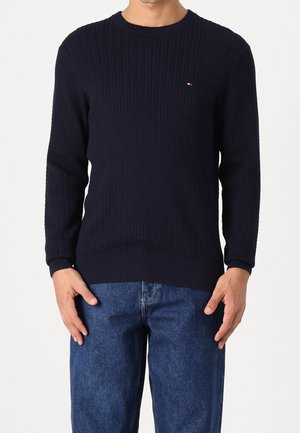 Mand iført en navyblå kabelstrikket trøje med et lille logo på brystet og blå denimjeans, stående med hænderne nær lommerne.