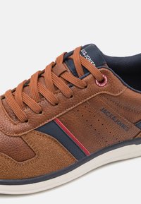 Hnědé kožené tenisky s texturovanými akcenty, námořnickými modrými pruhy, perforovanými detaily a oranžovými tkaničkami. Mají bílou gumovou podrážku a logo.