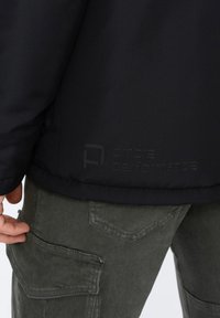 Černá péřová bunda s hladkým povrchem, která má decentní embosované logo "ombre performance" na dolní části zad, doplněná olivově zelenými cargo kalhotami.