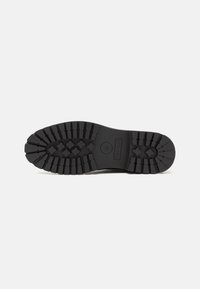 Suela de zapato de goma negra con profundas ranuras para tracción, que presenta un logotipo circular central y un diseño elegante y plano. Superficie texturizada para un mejor agarre.