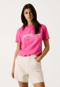 Rosa Baumwoll-T-Shirt mit weißem "Red Bull Racing"-Logo, kombiniert mit beigen Shorts mit fransigem Saum und seitlichen Taschen.