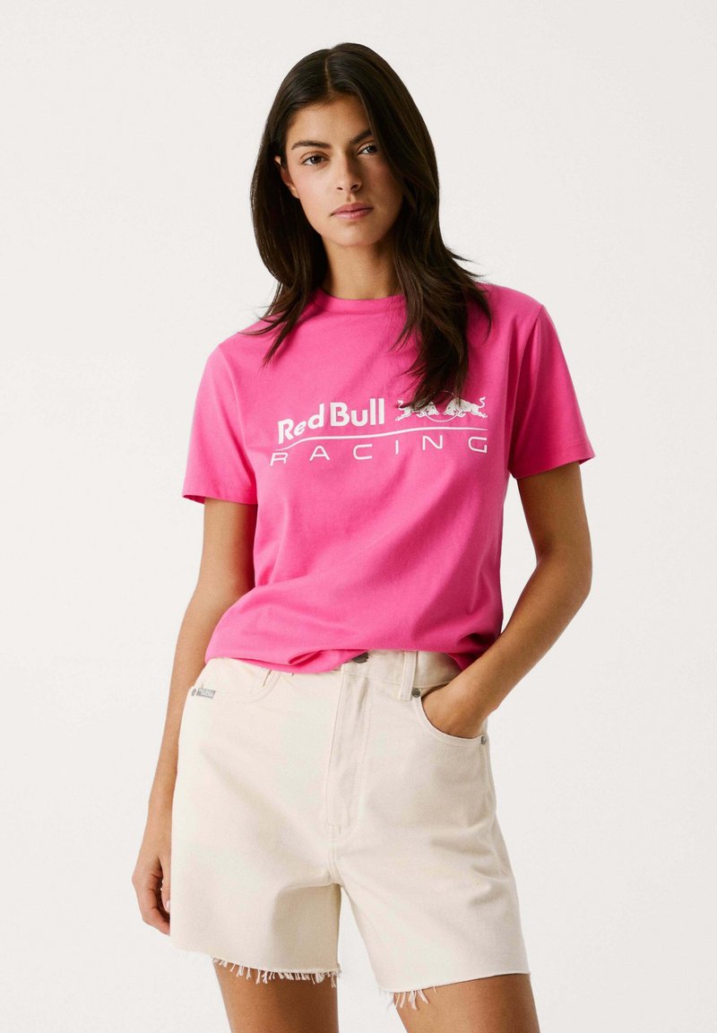 Camiseta de algodón rosa con el logotipo de "Red Bull Racing" en blanco, combinada con pantalones cortos beige con dobladillo deshilachado y bolsillos laterales.
