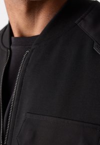 Veste bomber noire à texture lisse, fermeture éclair frontale et détails de poche. Design sans col ; coutures accentuées visibles.