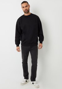 Zwarte sweatshirt met geribbelde boorden en onderkant, gecombineerd met donkere jeans en witte sneakers met zwarte accenten. Eenvoudige, casual outfit.