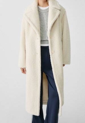 Person trägt einen langen, cremefarbenen Teddy-Mantel über einem grauen Cable-Knit-Pullover und marineblaue, weit geschnittene Hosen mit weißen Seitenstreifen.