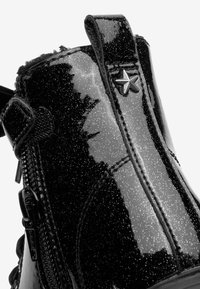 Musta lakattu nahkasaapas, jossa on kimaltava pinta, sivuvetoketju, tähden korostus ja vahvistetut saumat.