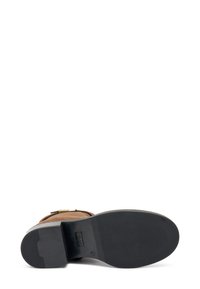 Stivale in suede marrone con suola texture, dotato di un accento in hardware dorato e un tacco piatto e liscio. Il design è alto alla caviglia.