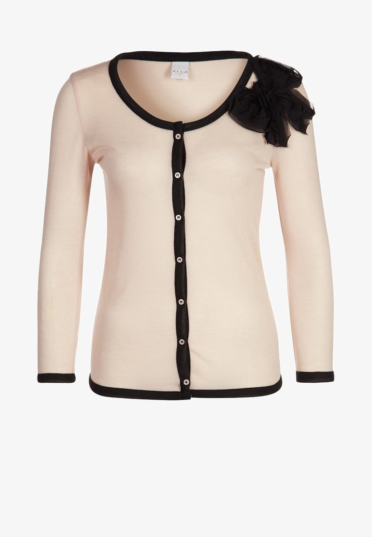 Cardigan rose pâle à manches longues avec bordure noire, boutons noirs sur le devant et une grande fleur en tissu noir sur l'épaule gauche.