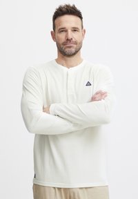 Langermet hvit henley-skjorte laget av ribbet stoff, med knappet hals, et lite trekantet logo på brystet, og syede mansjetter.