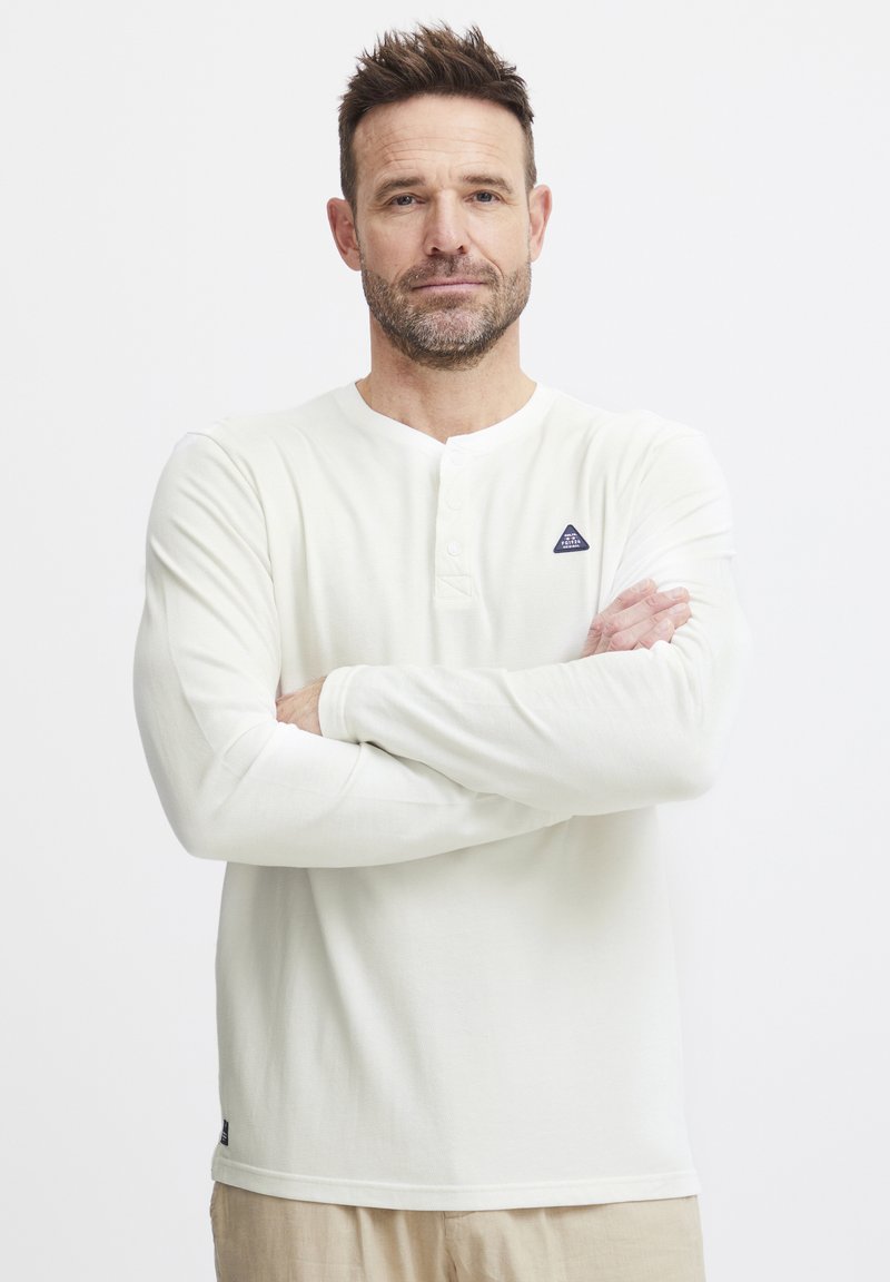 Langermet hvit henley-skjorte laget av ribbet stoff, med knappet hals, et lite trekantet logo på brystet, og syede mansjetter.