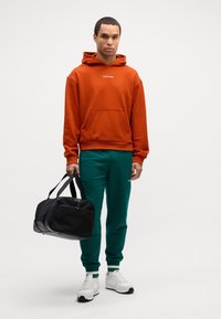 Felpa con cappuccio arancione e tasca frontale, abbinata a pantaloni della tuta verdi. Una borsa a tracolla nera con cerniera e scarpe da tennis bianche completano il look.