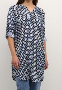 Camicia tunica blu navy con un motivo geometrico in azzurro chiaro. Presenta un collo a V, bottoni e maniche arrotolate; realizzata in tessuto leggero.