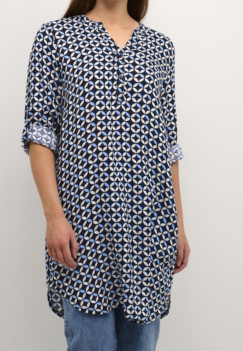 Camicia tunica blu navy con un motivo geometrico in azzurro chiaro. Presenta un collo a V, bottoni e maniche arrotolate; realizzata in tessuto leggero.