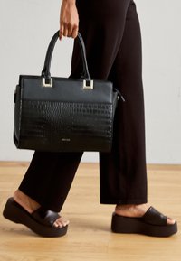 Sac à main noir structuré avec une texture crocodile, ornements en acier argenté. Associe à un pantalon noir évasé et des sandales à plateforme sombres.