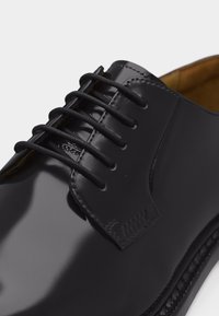 Fekete, fényesre polírozott bőrcipő zárt fűzővel és kerek fekete cipőfűzőkkel, fehér háttér előtt.