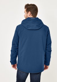 Mann in navyblauer Kapuzenjacke und dunklen Jeans, von hinten vor einem hellen Hintergrund betrachtet.