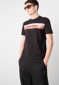 Svart bomull T-skjorte med en gradient rosa og hvit "Calvin Klein" logo på brystet. Kombinert med svarte bukser og brune solbriller.
