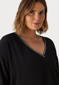 Top nera a scollo a V con rifinitura a strisce bianche e nere, caratterizzato da maniche corte e una texture morbida e liscia. Accentuato da una collana in catena dorata.