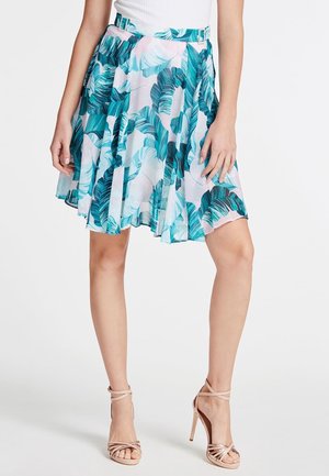 Femme portant une jupe fluide mi-longue avec un imprimé de feuilles tropicales vertes et bleu sarcelle, associée à des sandales à talons hauts couleur nude et un haut blanc.