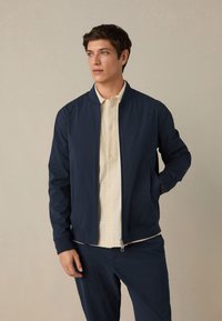 Jaunas vyras dėvi tamsiai mėlyną striukę su užtrauktuku ir atitinkančias kelnes, po kuria yra šviesaus kreminio atspalvio tekstūruota marškinėlių su sagomis, stovintis prie vientisos spalvos fono.