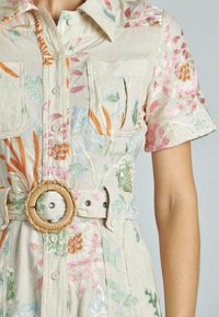 Leinenkleid mit buntem Blumenstickerei, kurzen Ärmeln, vorderen Taschen und einem gewebten Gürtel-Detail in der Taille. Heller beiger Hintergrund.