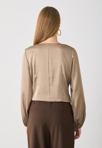 Korte beige satijnen blouse met lange mouwen en een ronde halslijn. Gladde textuur en subtiele achterkantnaad. Gecombineerd met bruine broeken.