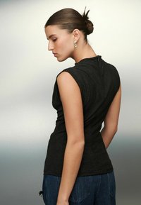 Noor, kelle juuksed on kõrgel bun'is, kannab hõbedasi kõrvarõngaid, seljas must ilma varrukateta top ja sinised teksad, seistes külgsuunas lihtsa gradine tausta ees.