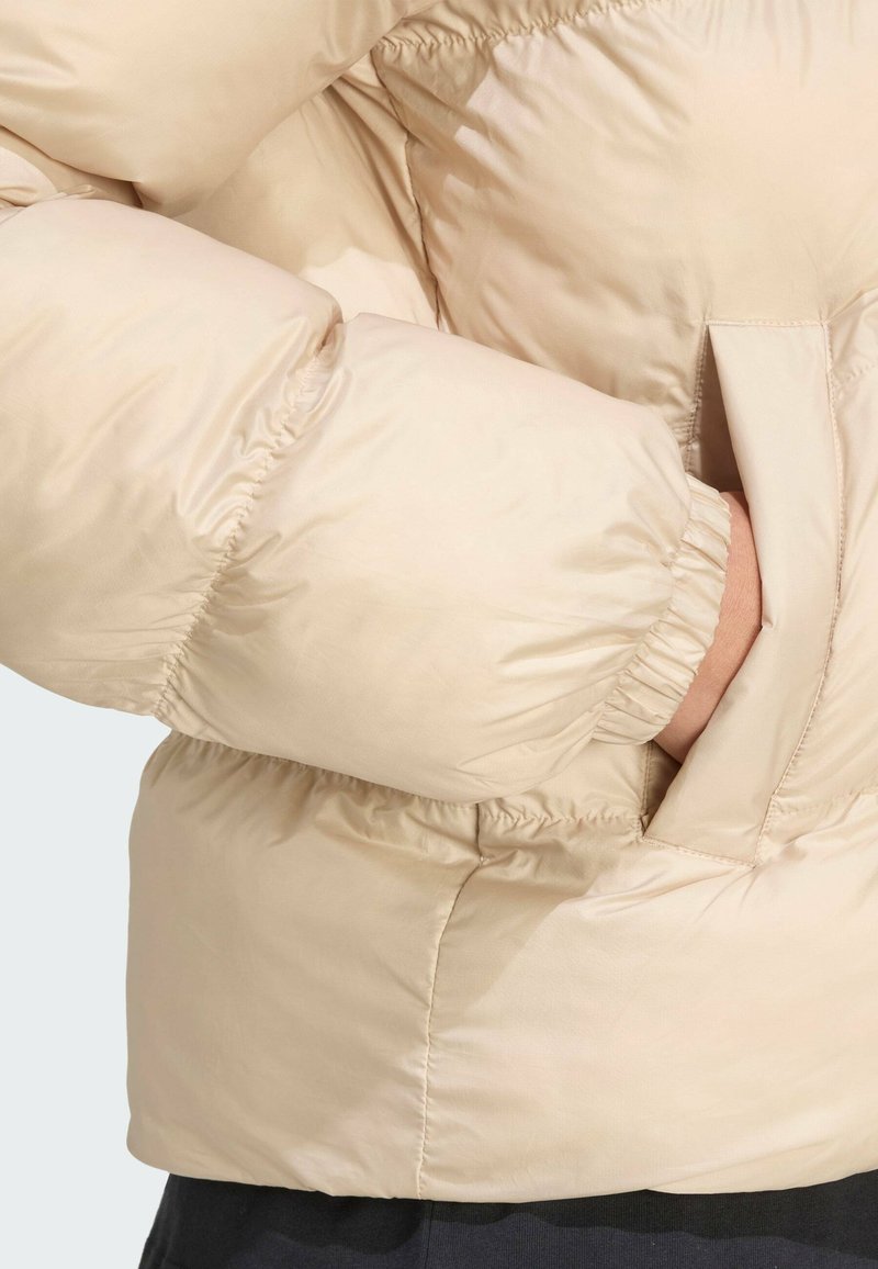 Giacca a piumino beige con superficie testurizzata, polsini elastici e tasche laterali. Il tessuto è liscio e leggermente lucido.