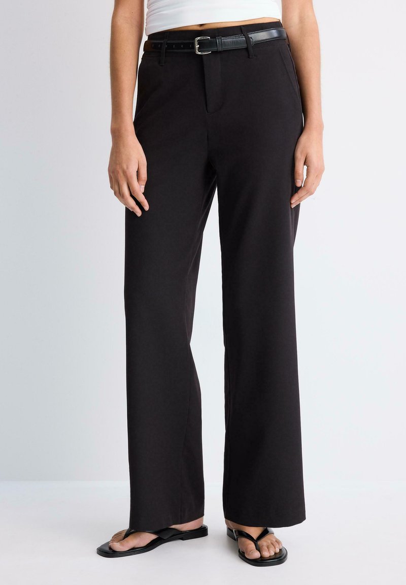 Femme portant un pantalon large noir avec une ceinture noire et des sandales noires, debout sur un fond uni.