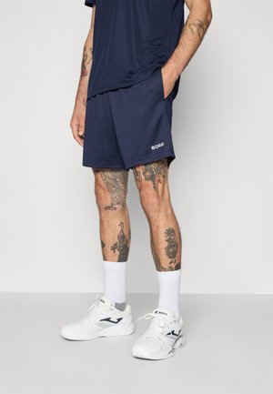 Man wearing navy shorts with "BORG" logo, white athletic sneakers with dark logo, and white socks, showing tattooed legs and arm.