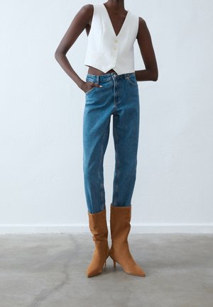 Personne mince portant un gilet blanc court boutonné, un jean taille haute bleu et des bottes marron à talons pointues et souples.