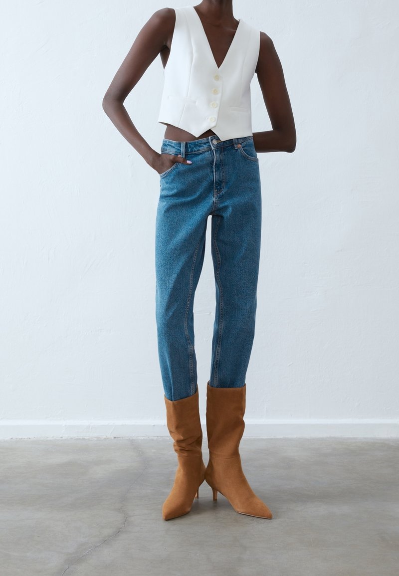 Personne mince portant un gilet blanc court boutonné, un jean taille haute bleu et des bottes marron à talons pointues et souples.